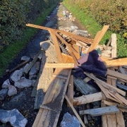 Wood planks, rocks, and debris block a narrow country lane with hedges on both sides.